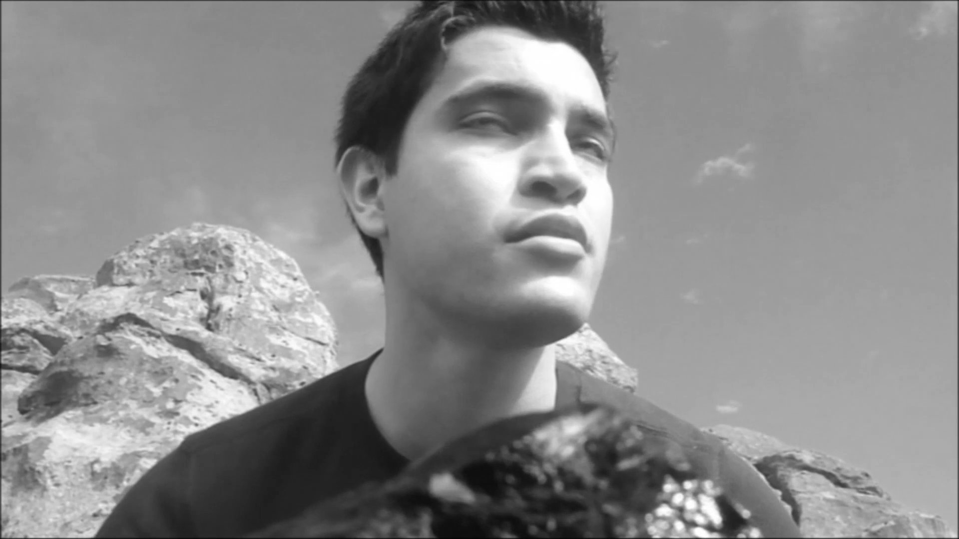 A man stands against a rugged rock formation, observing his surroundings with a thoughtful expression. The scene transitions from an abstract cosmic background to a focused personal portrait in an outdoor environment.