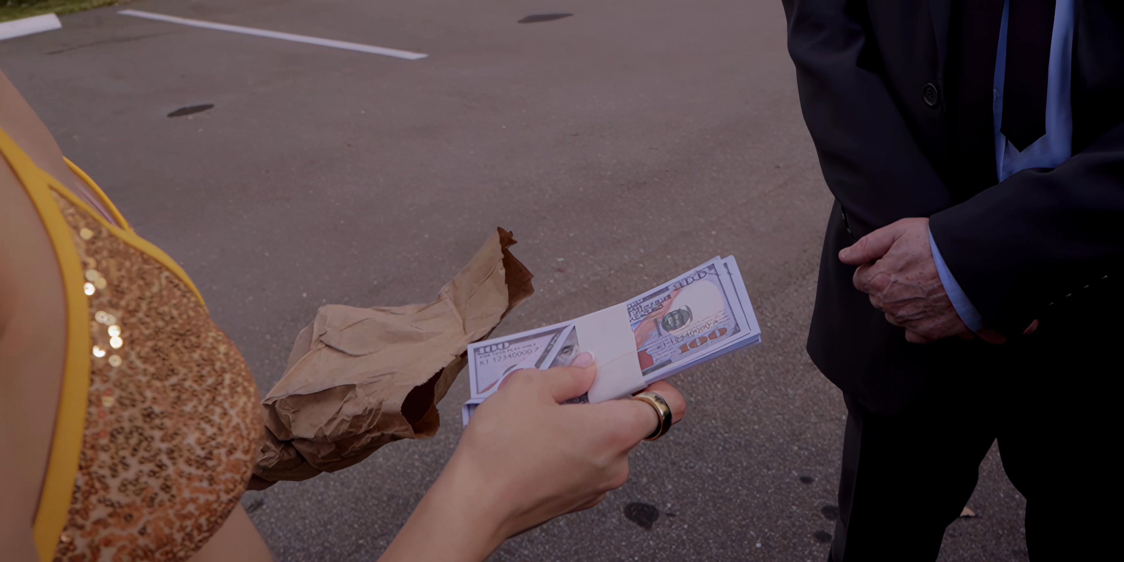 A man in a business suit engages in an exchange with a young woman in a parking lot, involving a paper bag and a sum of currency.