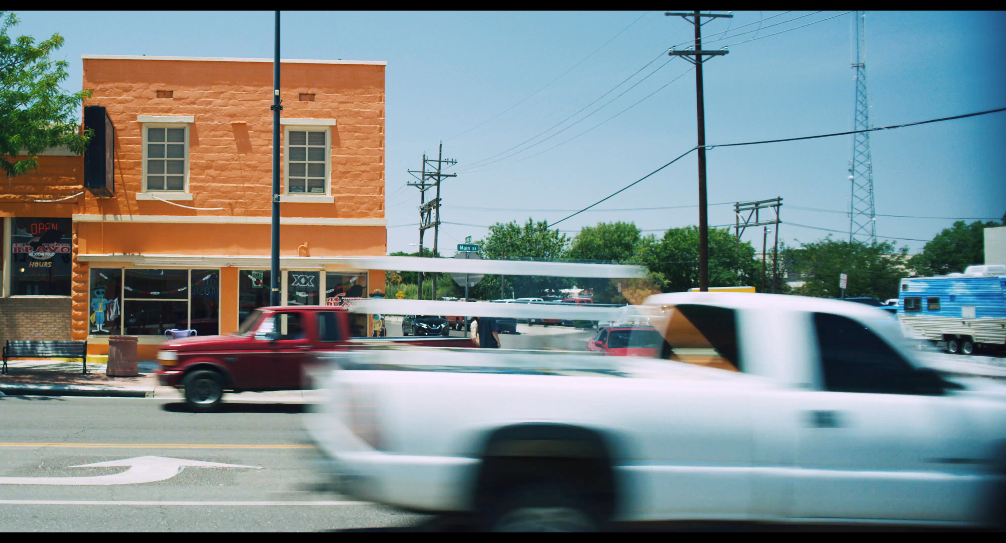 Two individuals walk along a city sidewalk while engaging in conversation on a sunny day. The scene transitions between their movement along building storefronts and the surrounding traffic on the nearby street.