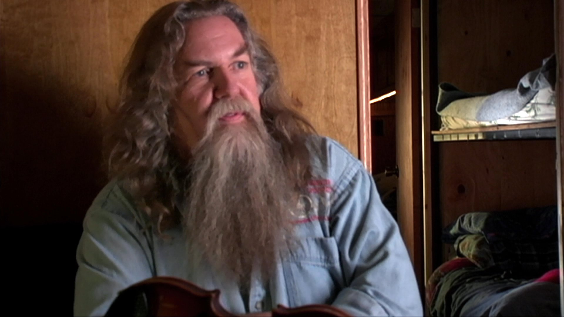 A long-haired man with a thick beard delivers an expressive monologue, using hand gestures to emphasize his storytelling. He sits before a wooden-paneled background while actively engaging with an unseen audience.