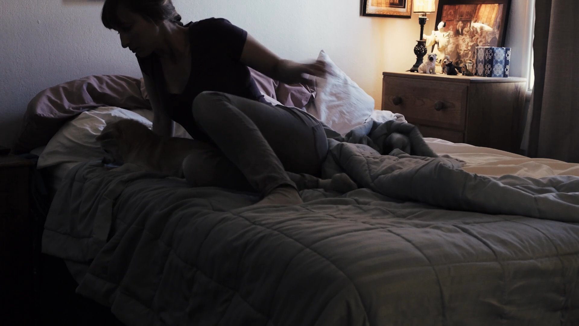 A woman wakes up in her bedroom and gently interacts with her small dog before starting her morning. She then prepares to move from the bed while the room transitions into daylight.