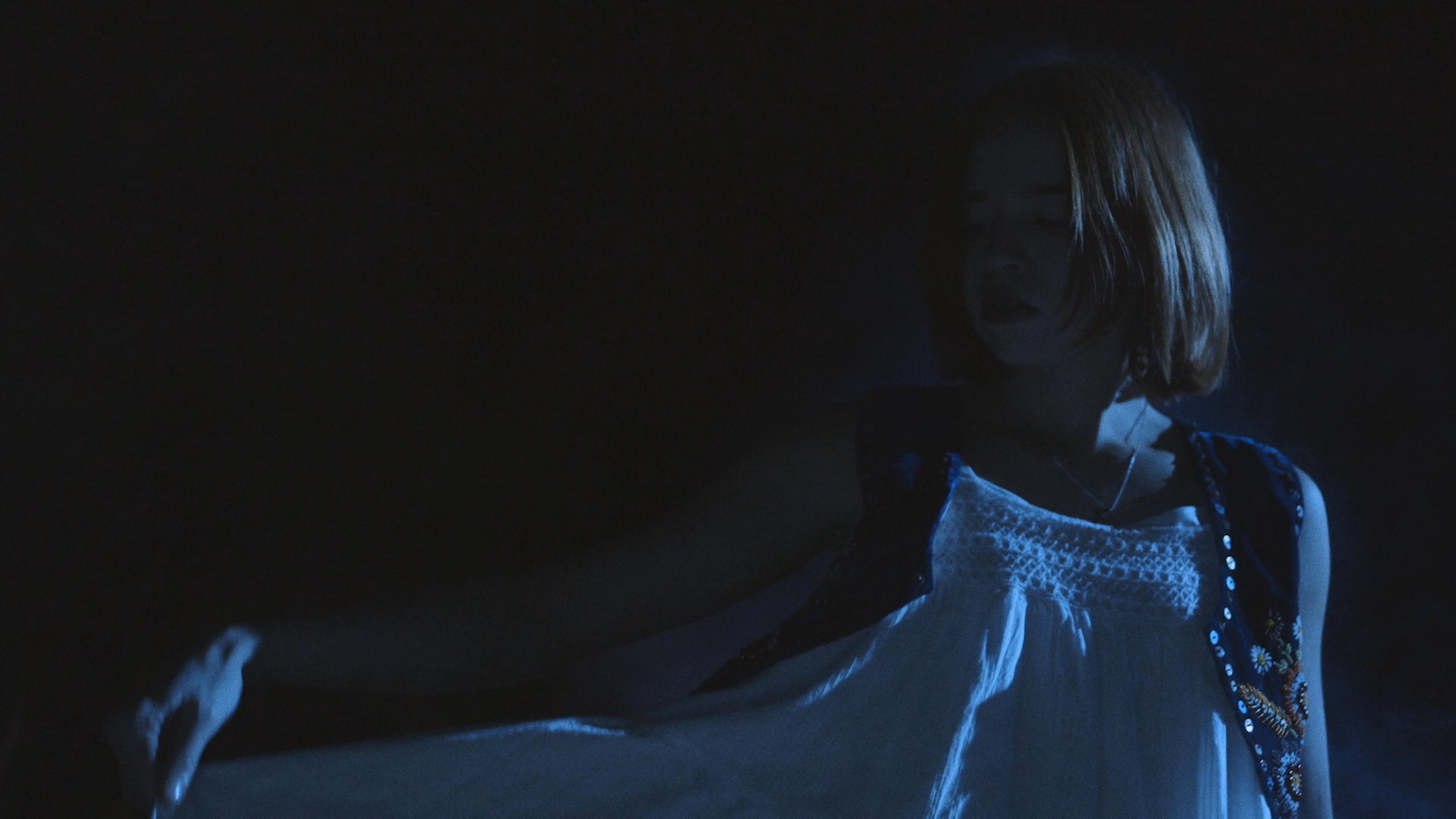 A woman in a white dress and vest performs a graceful dance in a dim, smoke-filled room illuminated by blue light. The atmospheric scene transitions between close-up shots of her movement and wide shots showcasing the expansive, darkened interior.