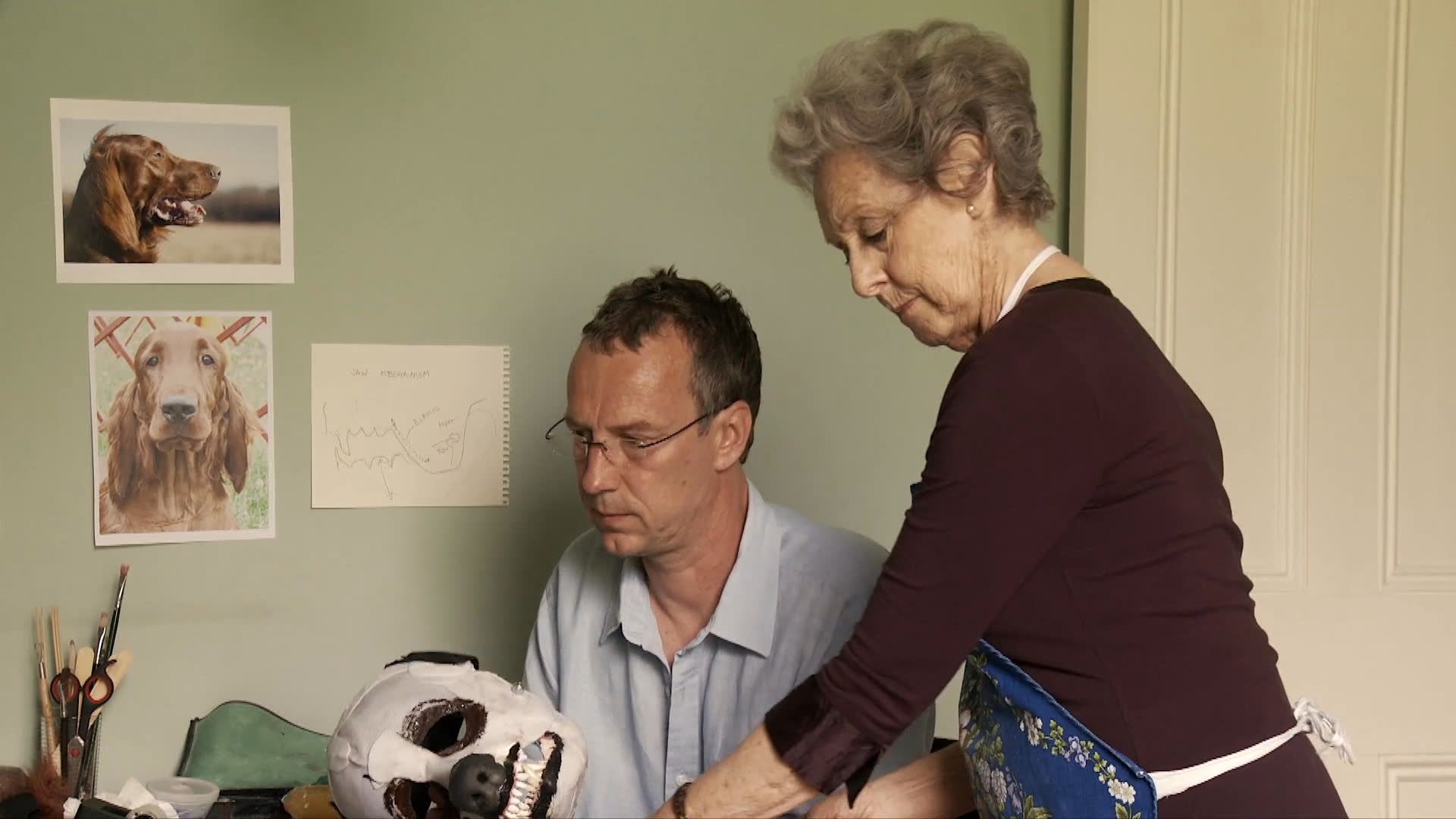 An experienced woman guides a male colleague in a detailed craft project involving a mask in a studio setting. They deliberate on design choices and coordinate their efforts to refine the work.