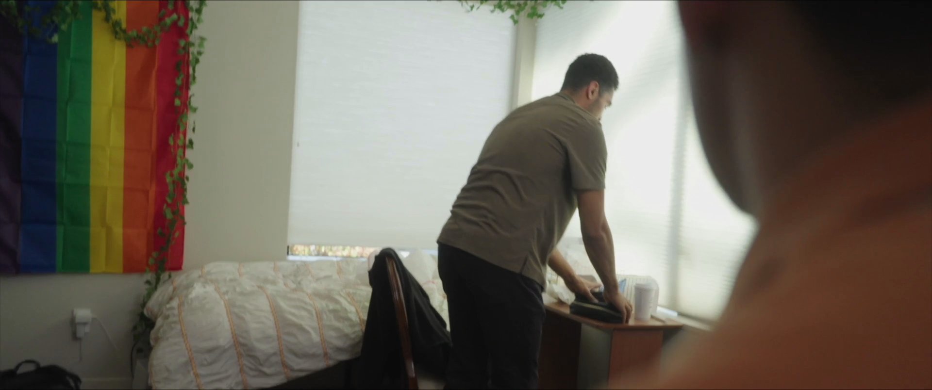 A man organizes personal items in a bedroom space while another individual observes from the background.