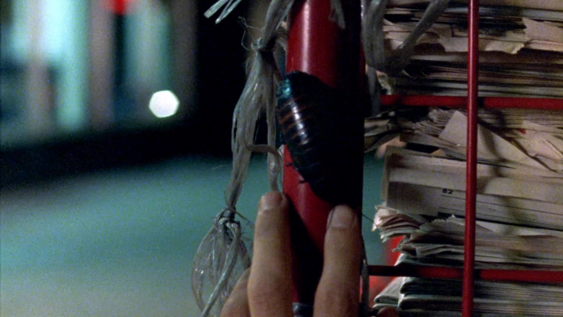 A man at an outdoor newspaper stand closely observes a large cockroach crawling along a metal display rack during the night. He watches the insect carefully as it moves across the structure.
