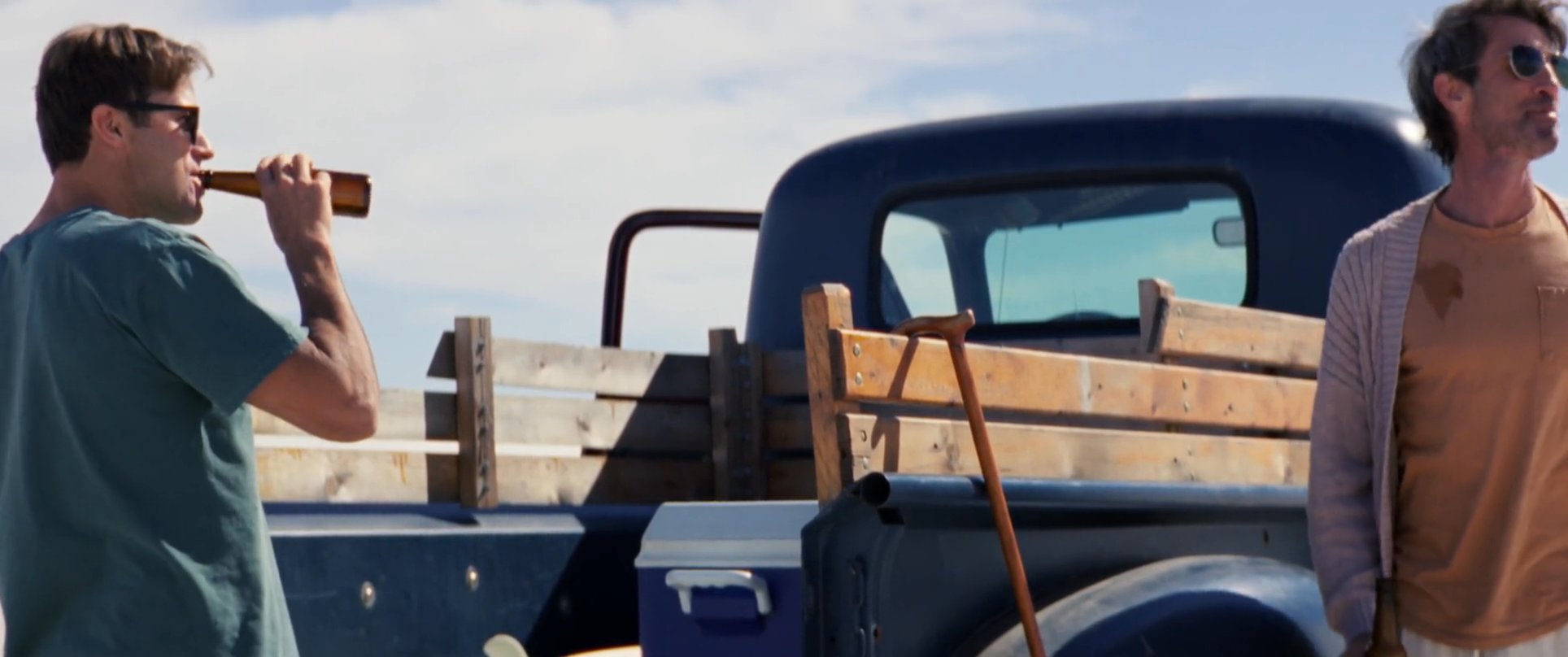 Two men enjoy drinks near a vintage pickup truck parked amidst vast, sunlit sand dunes. The pair engages in casual conversation while taking in the expansive desert landscape.