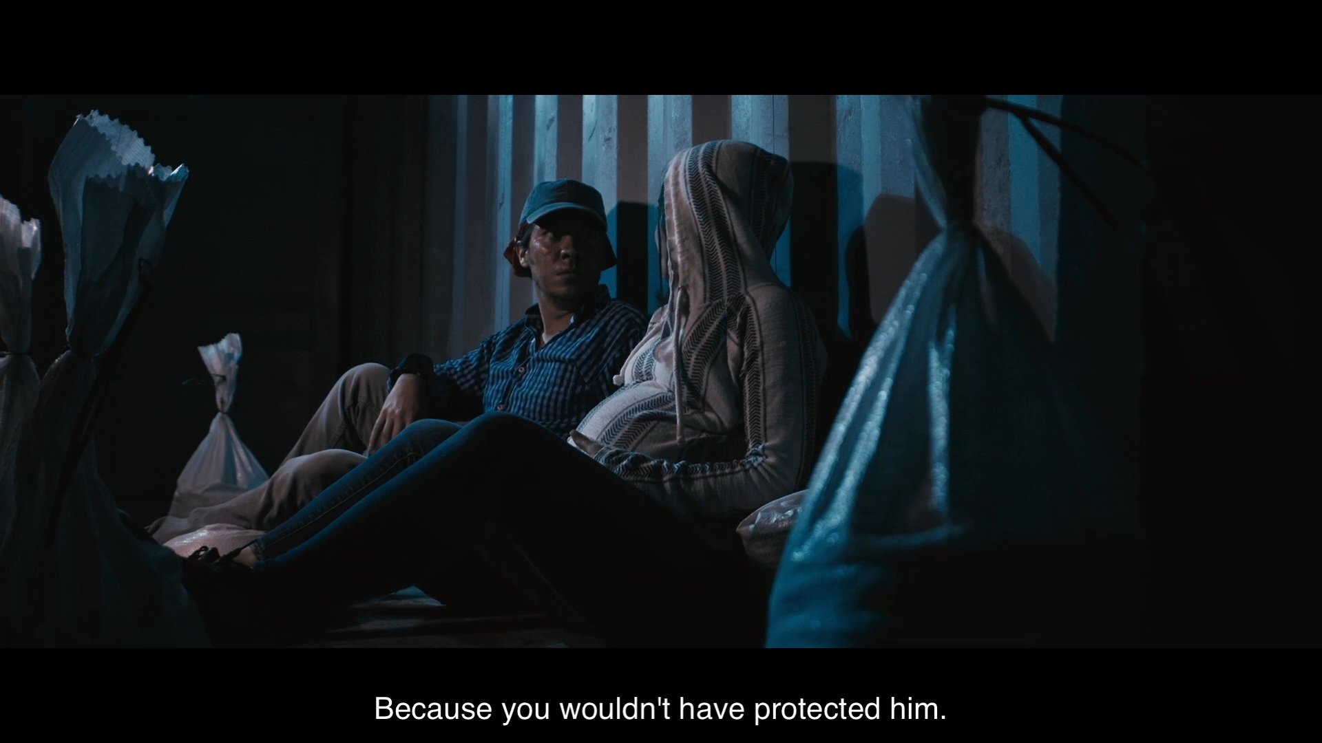A man and a woman sit in a dimly lit space, engaging in a tense dialogue about protection and past events. The mood remains somber as they reflect on an encounter that was narrowly avoided.