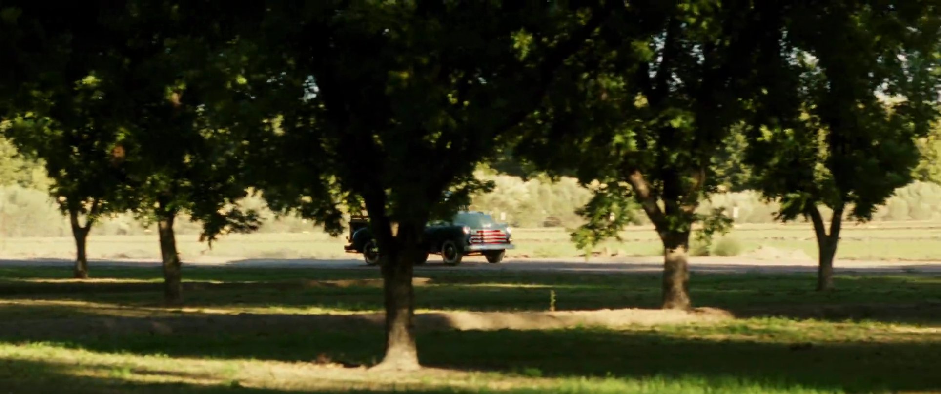 A man driving a vintage pickup truck arrives at a rural farmstead during the golden hour, navigating through tree-lined paths towards a white farmhouse.