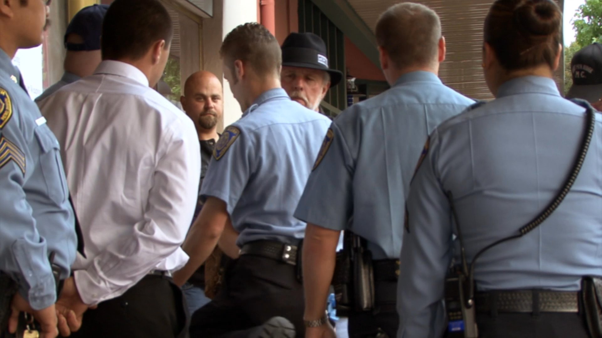 Members of a motorcycle club engage in a tense, supervised interaction with police officers outside of a retail establishment. The scene captures the visual contrast between the club members' apparel and the uniformed authorities during an ongoing investigation or inquiry.