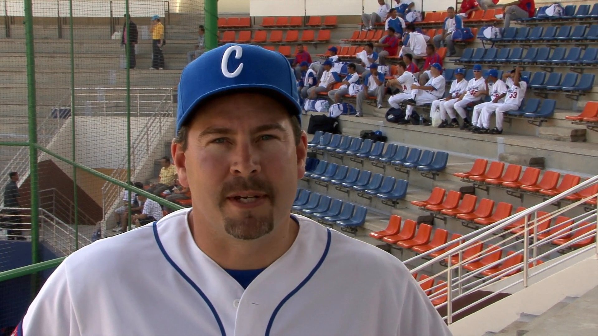Two individuals wearing baseball uniforms discuss the game from within a stadium, interspersed with shots of evening city traffic.