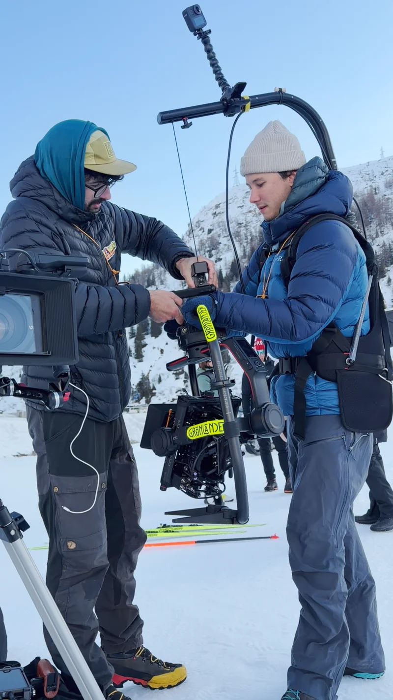 Two crew members adjust a body-mounted camera rig during a snowy mountain shoot.