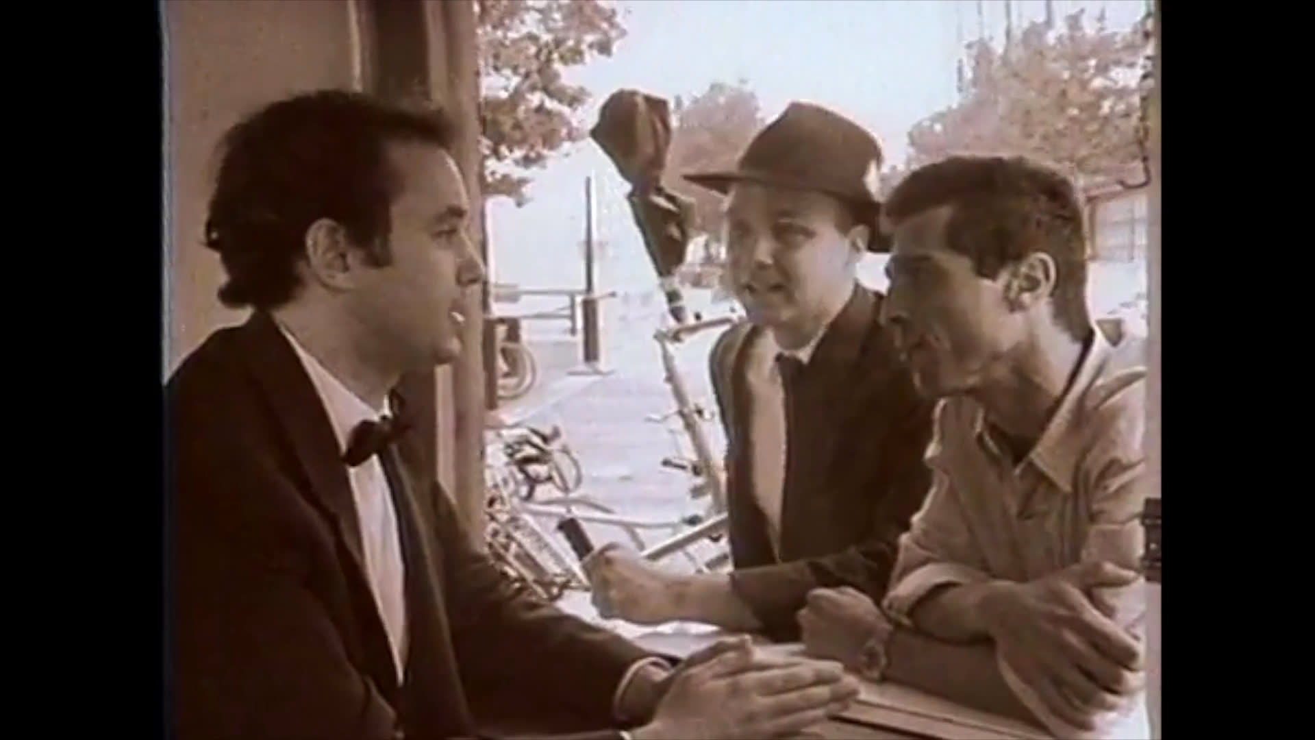 Three men, including two dressed in suits and hats, engage in an earnest conversation while sitting at an outdoor table in a sepia-toned setting.