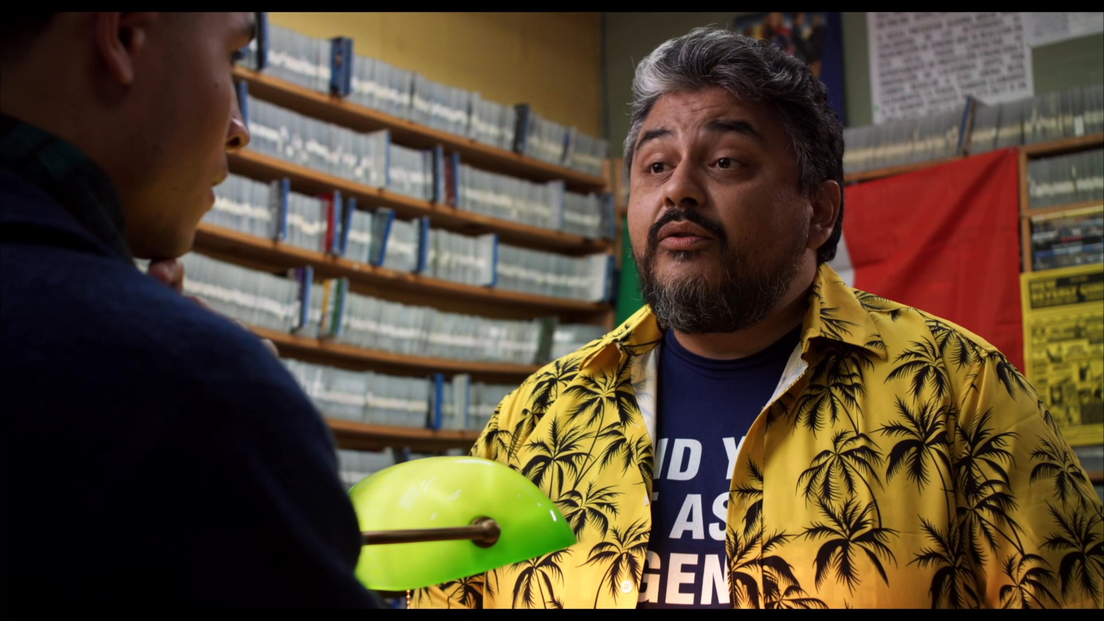 A customer and a store clerk engage in a steady conversation behind a counter filled with movie inventory in a video rental shop.