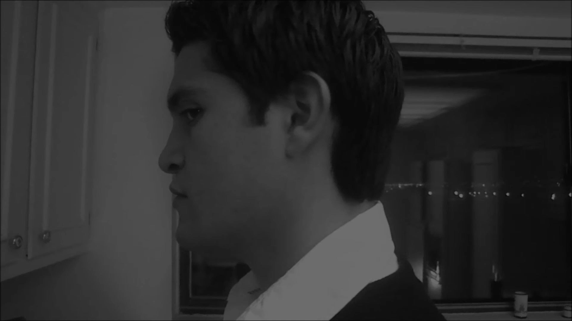A man paces anxiously in a dim kitchen before stopping to look directly into the camera with an intense expression. He then walks toward a nearby table, reaching for an object beside a framed photograph.