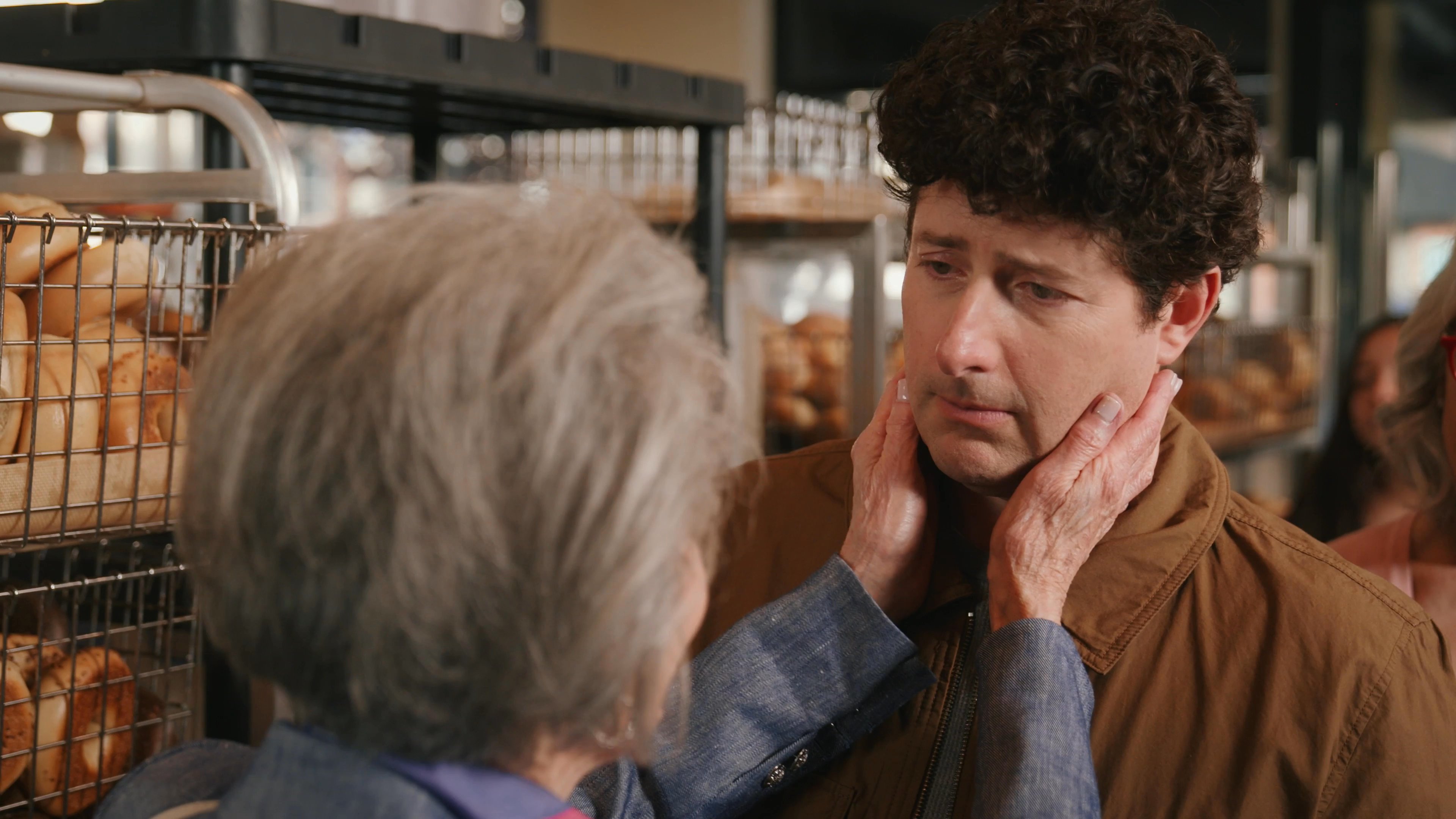 An older woman and a man share an intimate, emotional moment of connection and physical affection within a busy bakery.