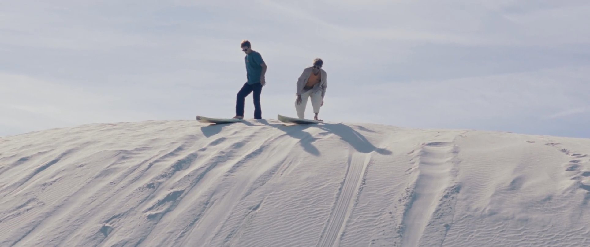 Two friends share an emotional embrace before sandboarding together down the face of a vast desert dune.