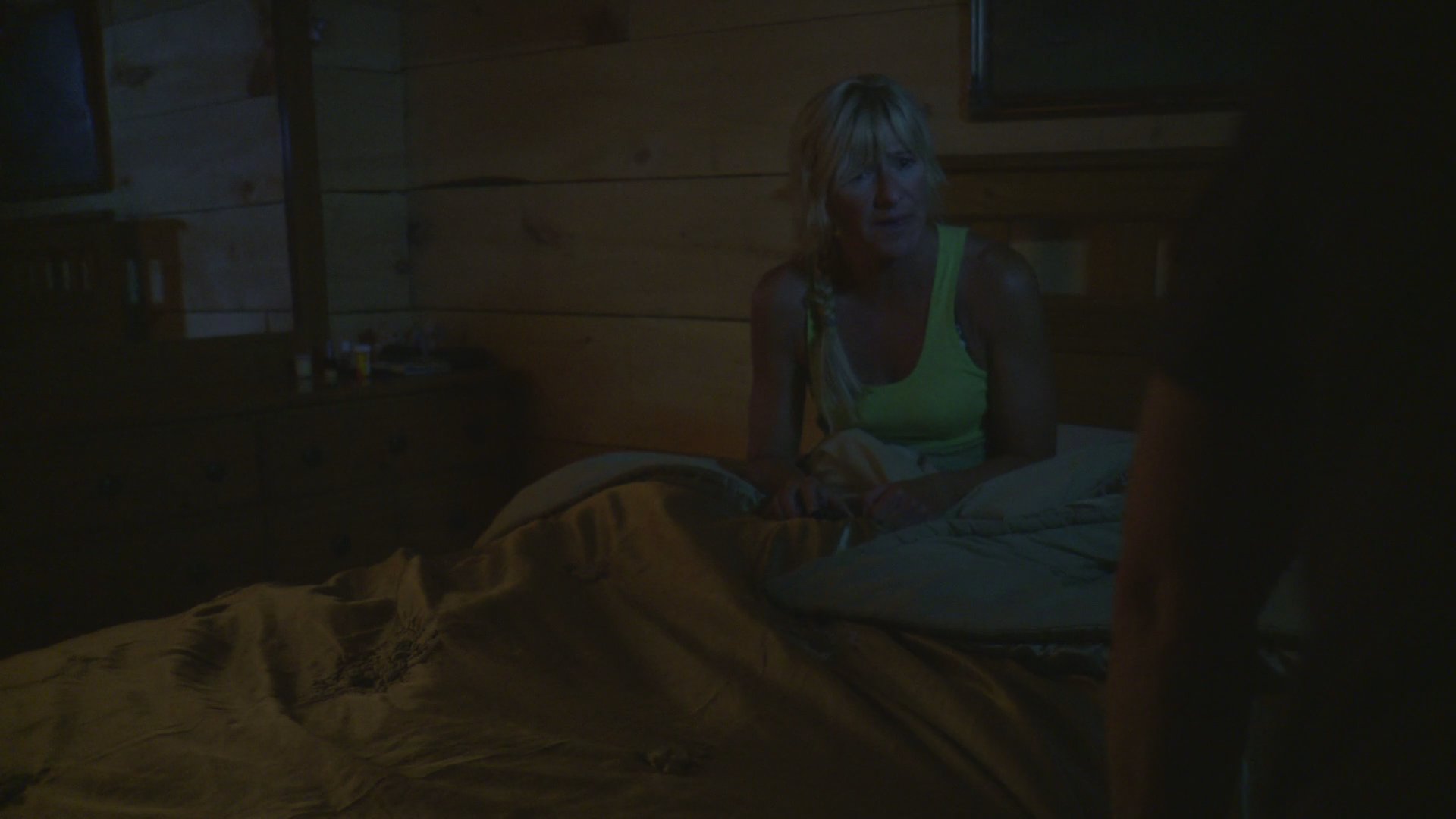 A man wakes up abruptly in bed during the night, visibly distressed, while his partner observes him in the dim, wooden-walled bedroom.