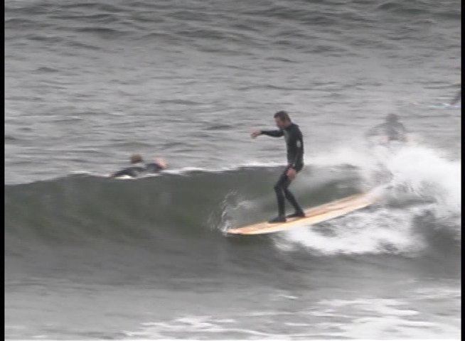 A surfer in a wetsuit skillfully executes a noseride maneuver on a longboard while traversing the face of a rolling ocean wave.