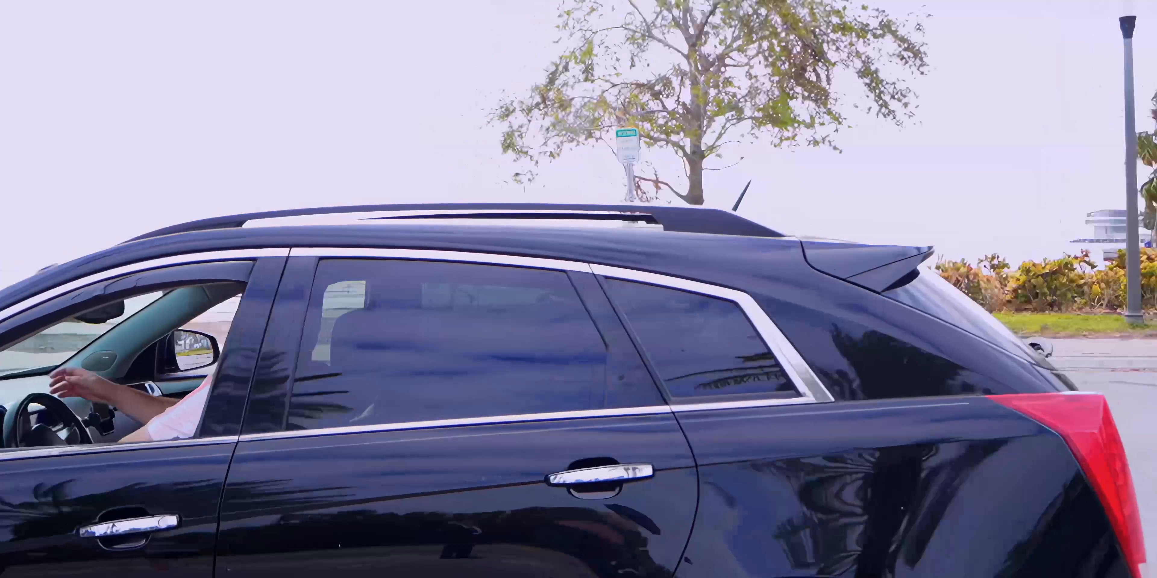 A dark SUV navigates a parking lot located near a coastline, eventually coming to a stop as a man walks along the nearby waterfront.