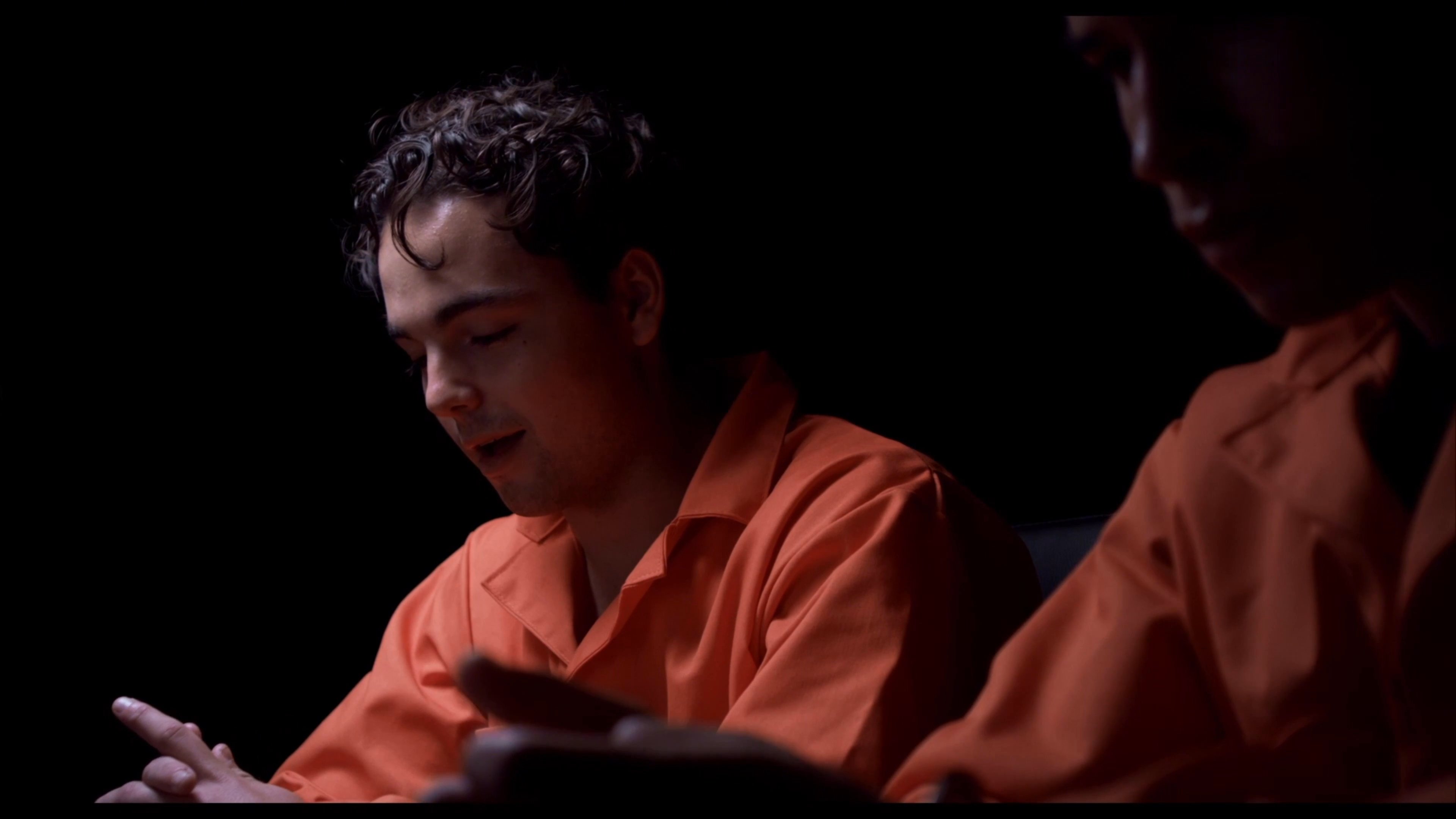 Two individuals dressed in orange prison jumpsuits engage in a tense, hushed conversation while handcuffed in a dark interrogation room. The dialogue unfolds with deliberate gestures, suggesting a high-stakes exchange between the detainees.