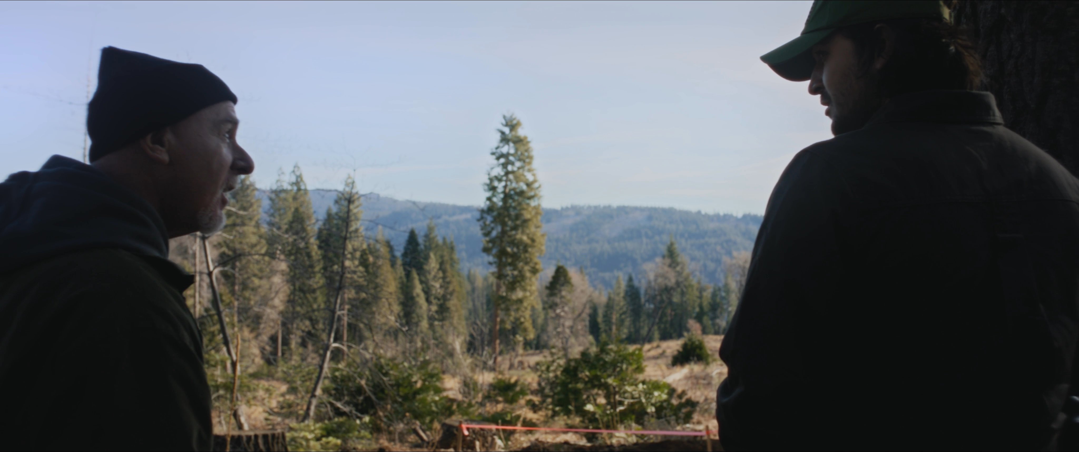 Two men, including a sheriff, confer at a remote forest location overlooking an excavation site. They examine the earth and surrounding terrain as part of an official inquiry.