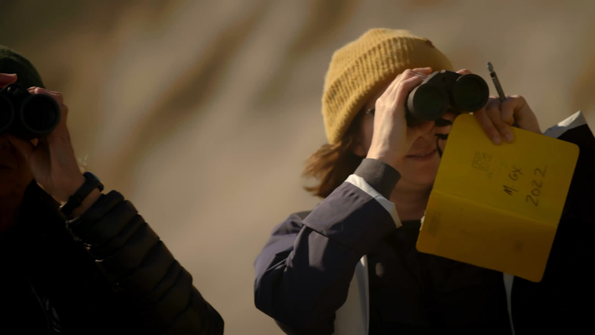 Three women scan a broad beach beneath steep coastal cliffs, using binoculars and field equipment to observe the shoreline. An experienced marine mammal researcher reflects on entering a male-dominated field in the 1970s.