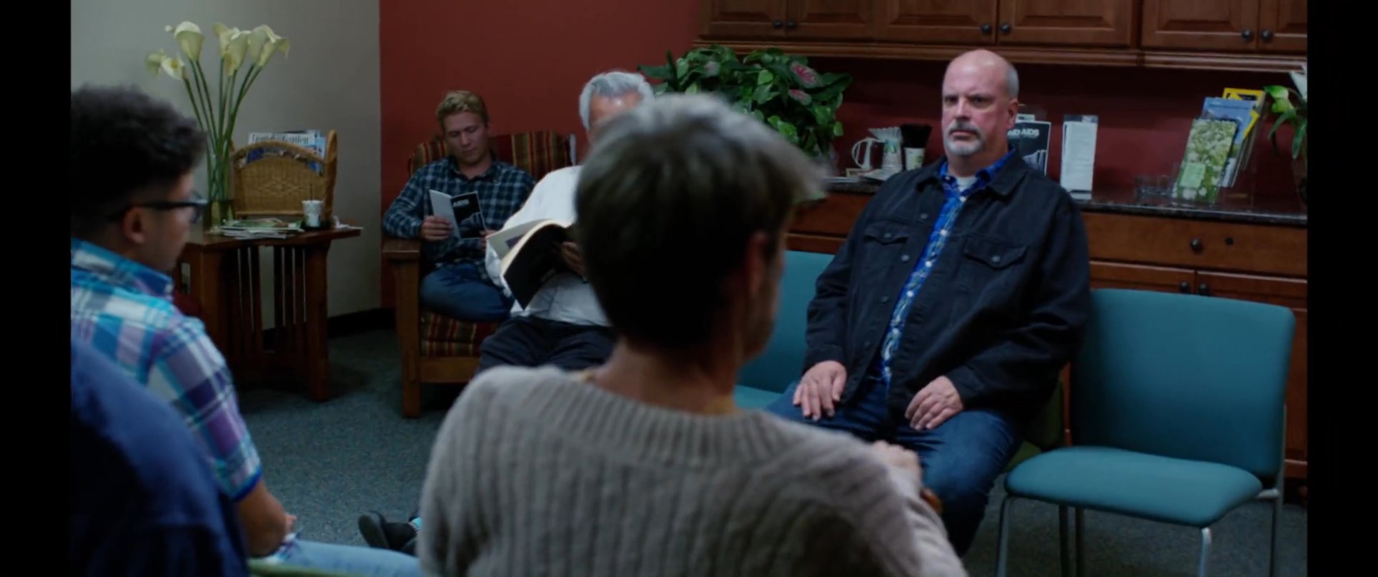 A man interacts with a receptionist before joining a group of people seated in a waiting area. The scene transitions to the man engaging in quiet observation among others in a professional, clinical setting.
