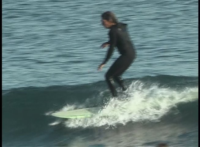 A surfer wearing a black wetsuit skillfully navigates a rolling ocean wave on a longboard. The individual maintains balance and control while gliding across the water surface.