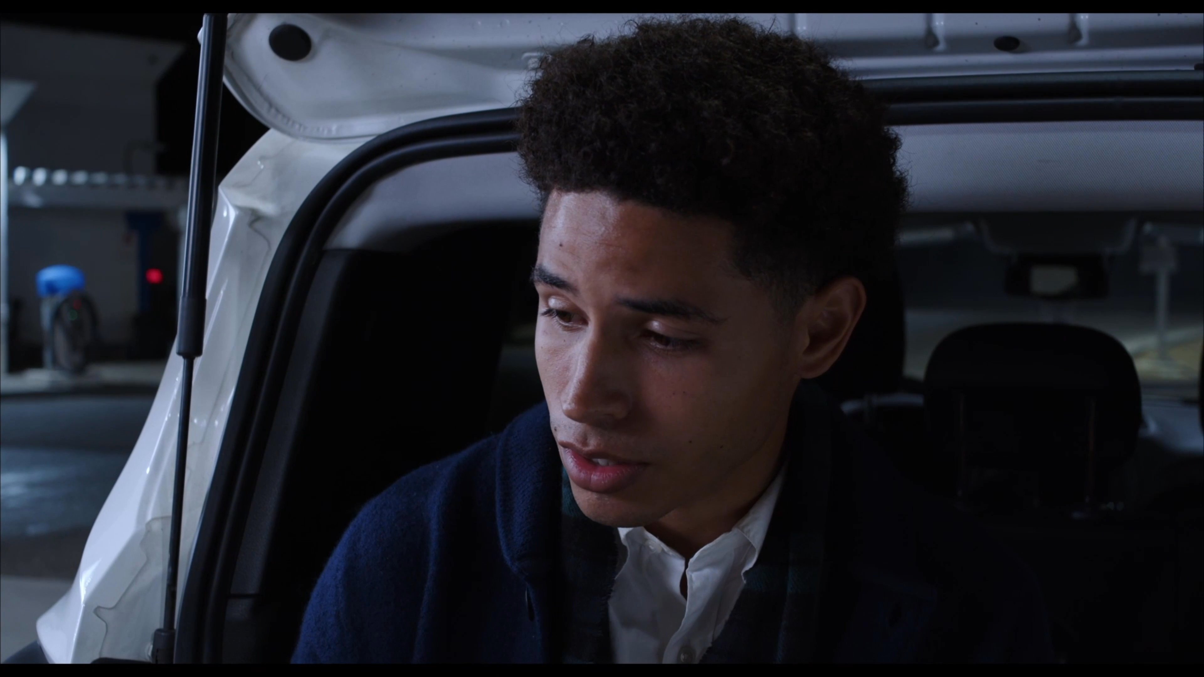 Two young men engage in a quiet conversation at a gas station at night, with one individual sharing a snack with the other near an open vehicle trunk.