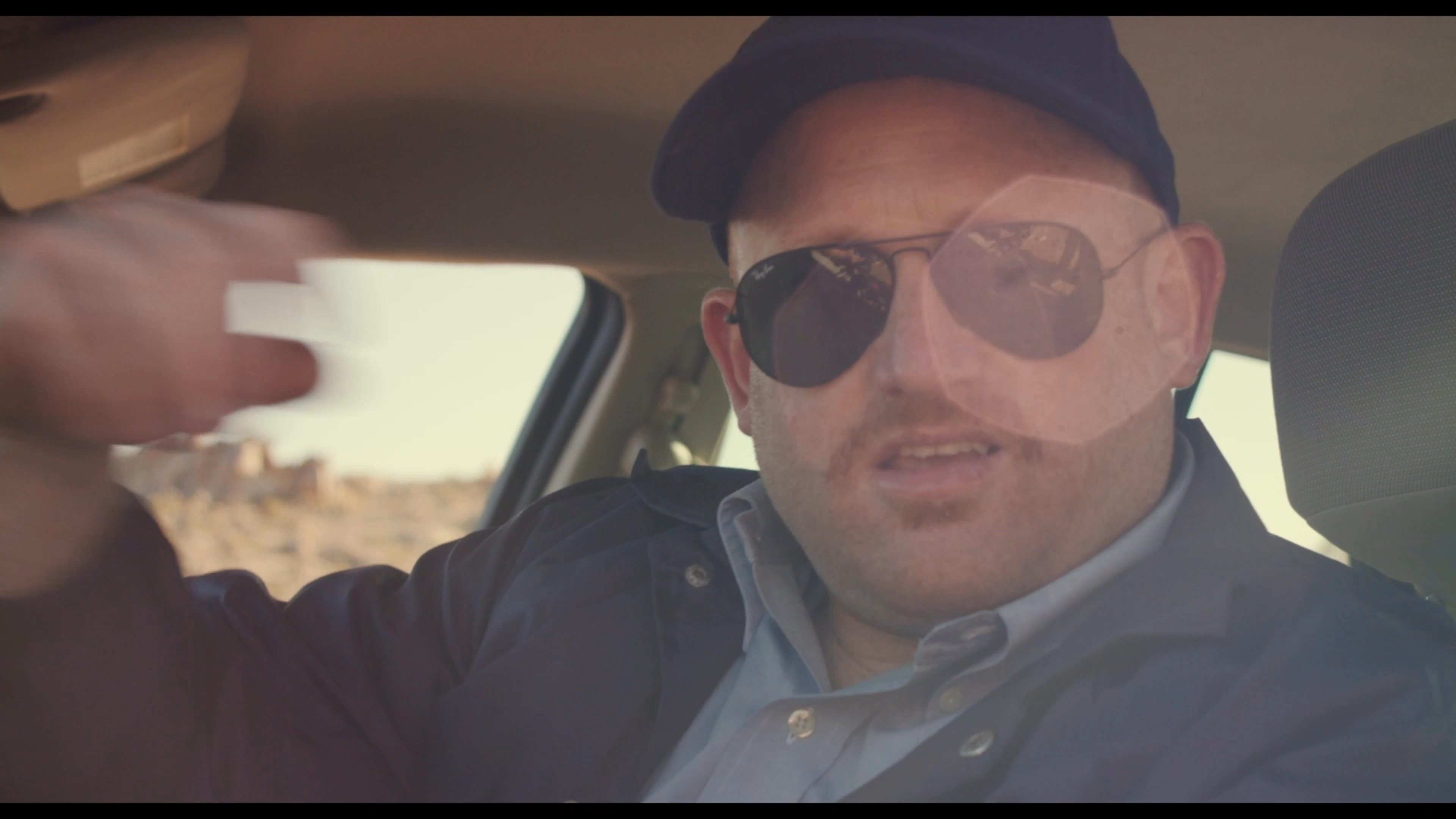A police officer monitors a mobile device while parked in a remote desert setting, later exiting his patrol car to utilize a two-way radio.