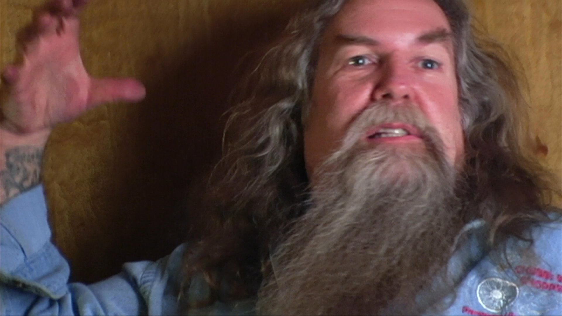 A long-haired, bearded musician reflects on his experiences while transitioning between candid storytelling and an intimate violin performance. He speaks directly to the camera before playing, framed against a poster featuring Willie Nelson.