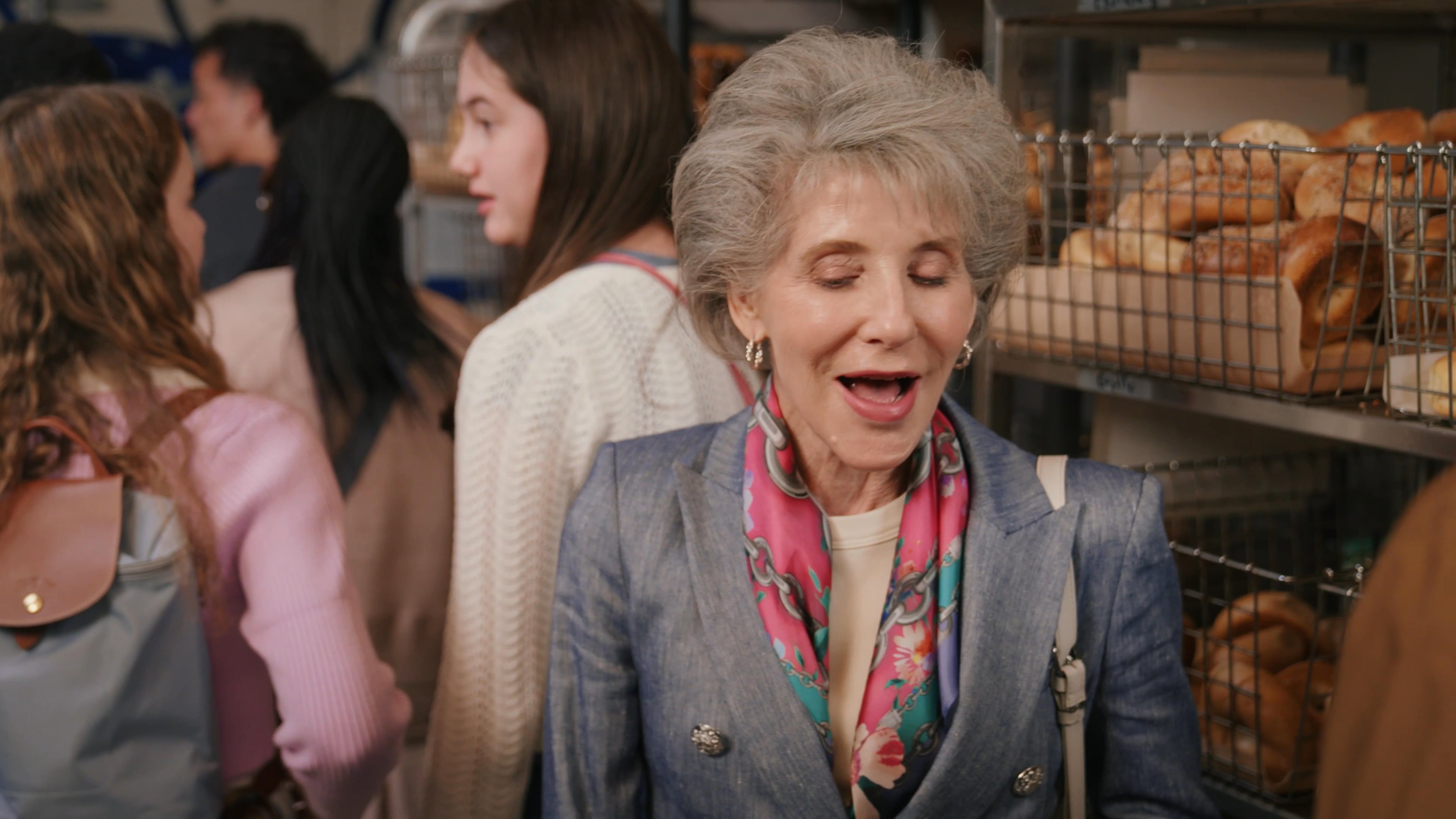 An older woman in a blue jacket engages in an animated conversation with a younger man while standing in front of shelves stocked with baked goods. They discuss the product display in a busy bakery setting.