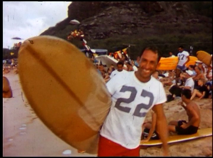 A surfer competes on large ocean waves, intercut with scenes of a beach competition and a reflective interview regarding the sport's history.