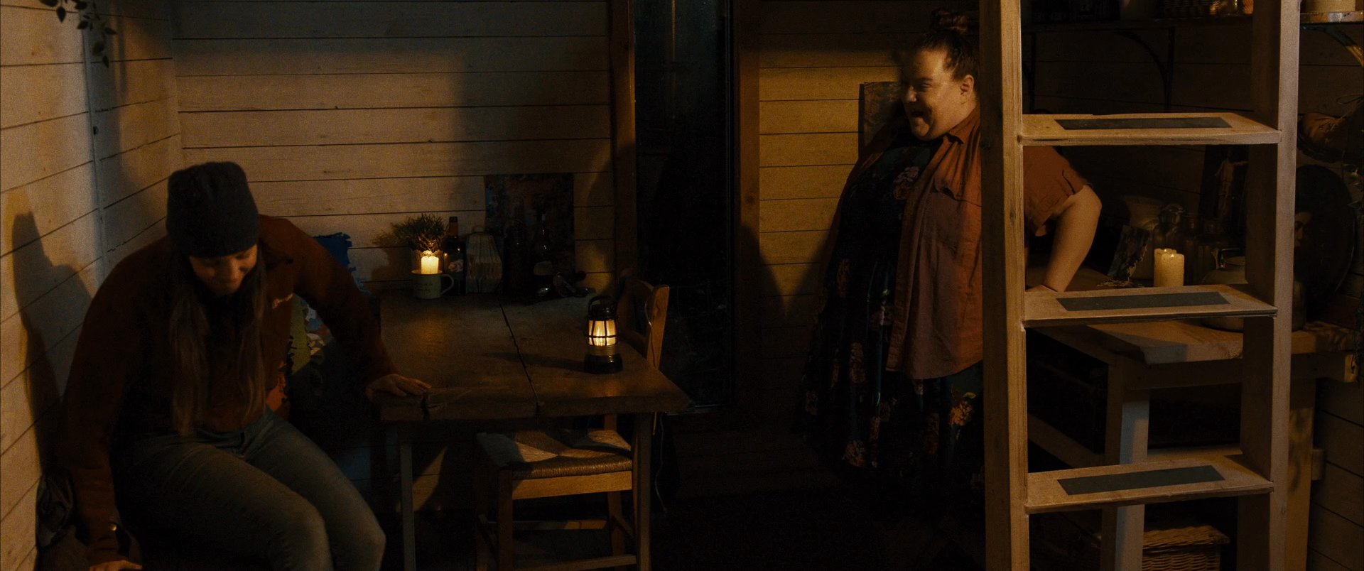 Two individuals engage in an intimate conversation within a dimly lit, rustic cabin environment. The pair interacts naturally while seated in a warm, low-light setting.