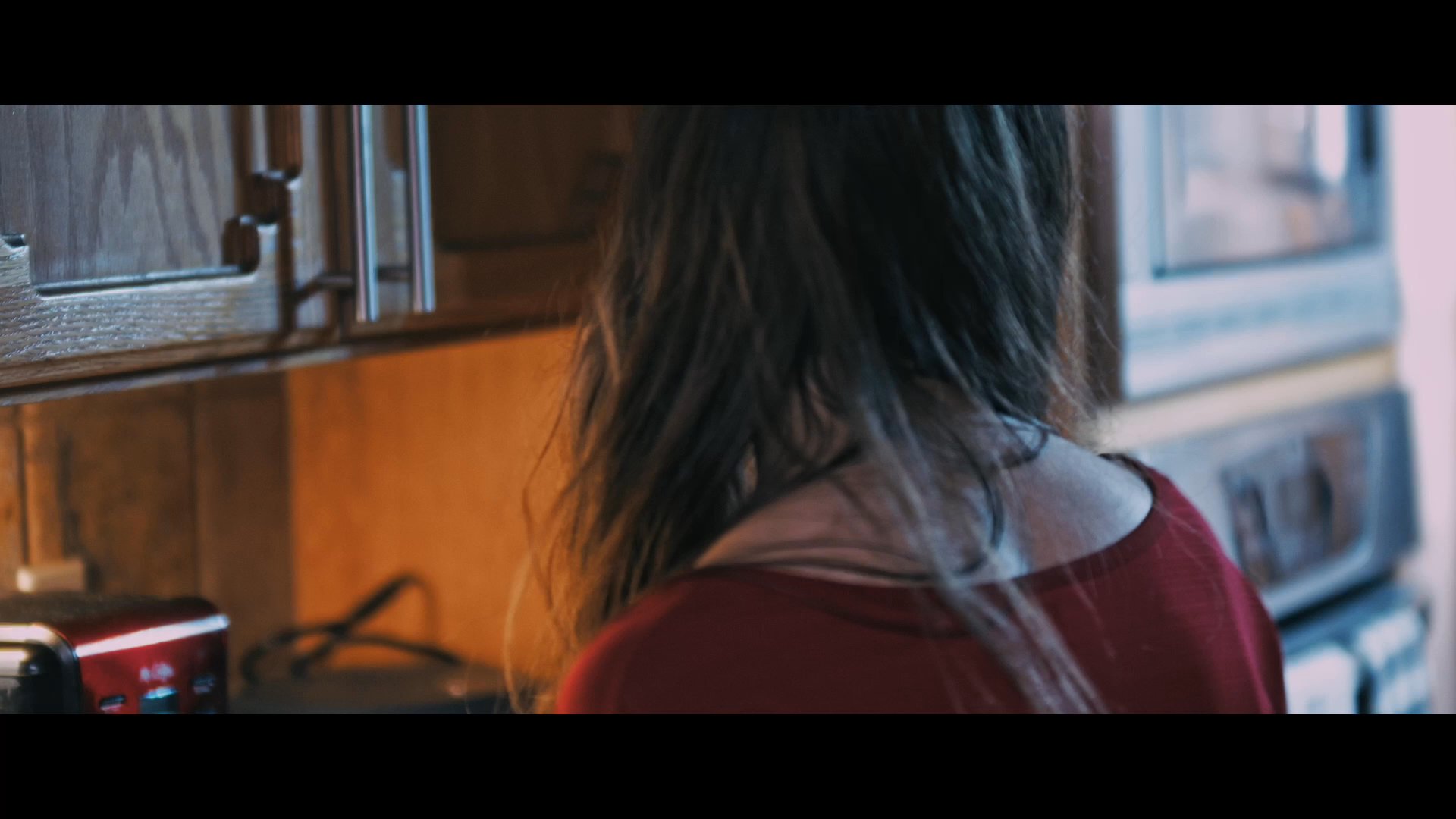 A woman stands in a dim kitchen interacting with her phone before turning to gaze out a window. Her demeanor reflects a state of quiet contemplation or anticipation.