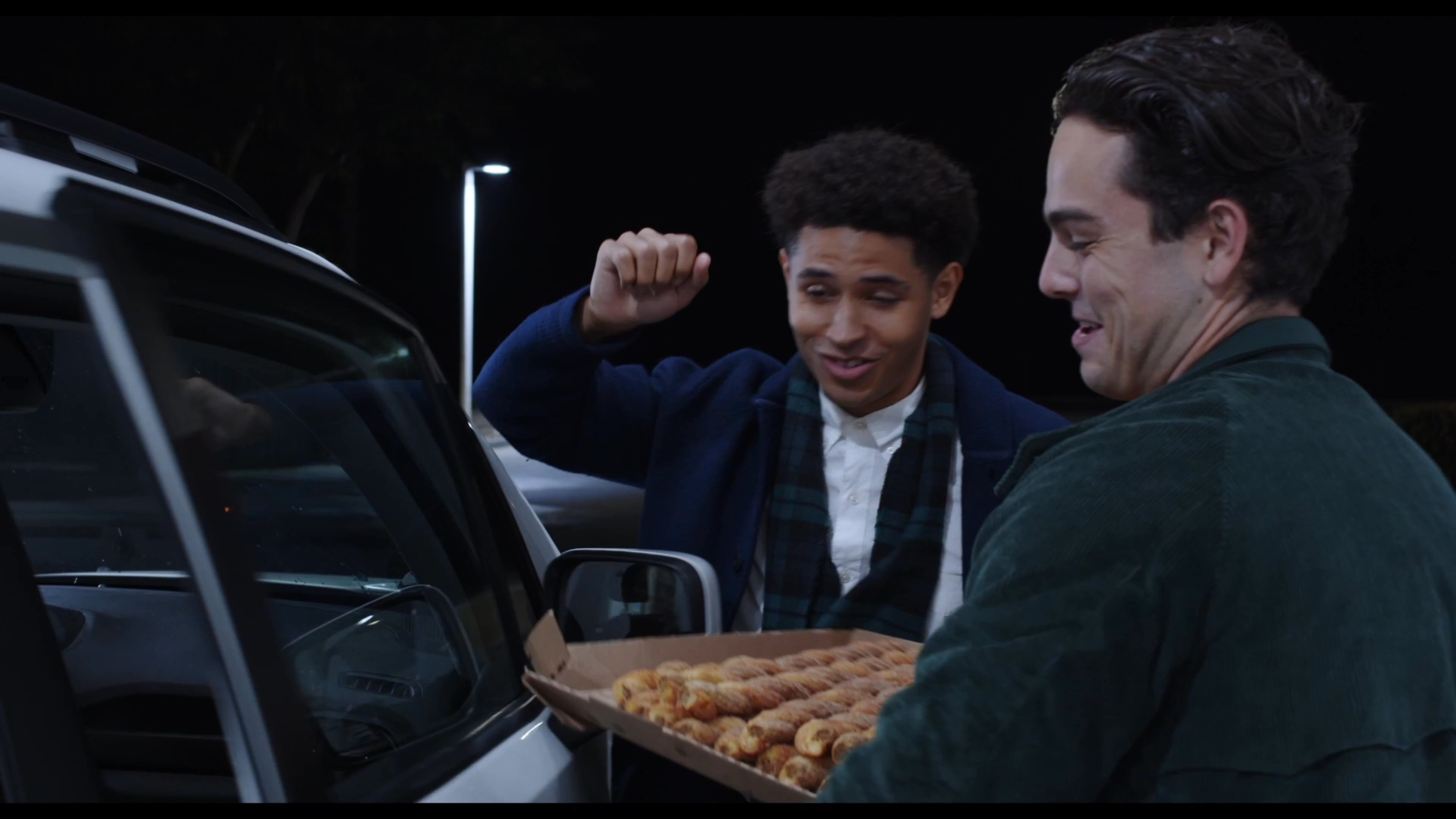 Two men engage in a friendly conversation next to a vehicle at night while one holds an open box of food. They maintain an relaxed demeanor during their exchange in an outdoor setting.