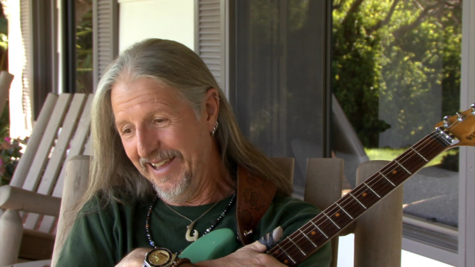 An experienced guitarist with long hair speaks directly to the camera while holding his instrument during an informal interview. He maintains an engaging presence as he discusses his musical background in a sunlit outdoor setting.