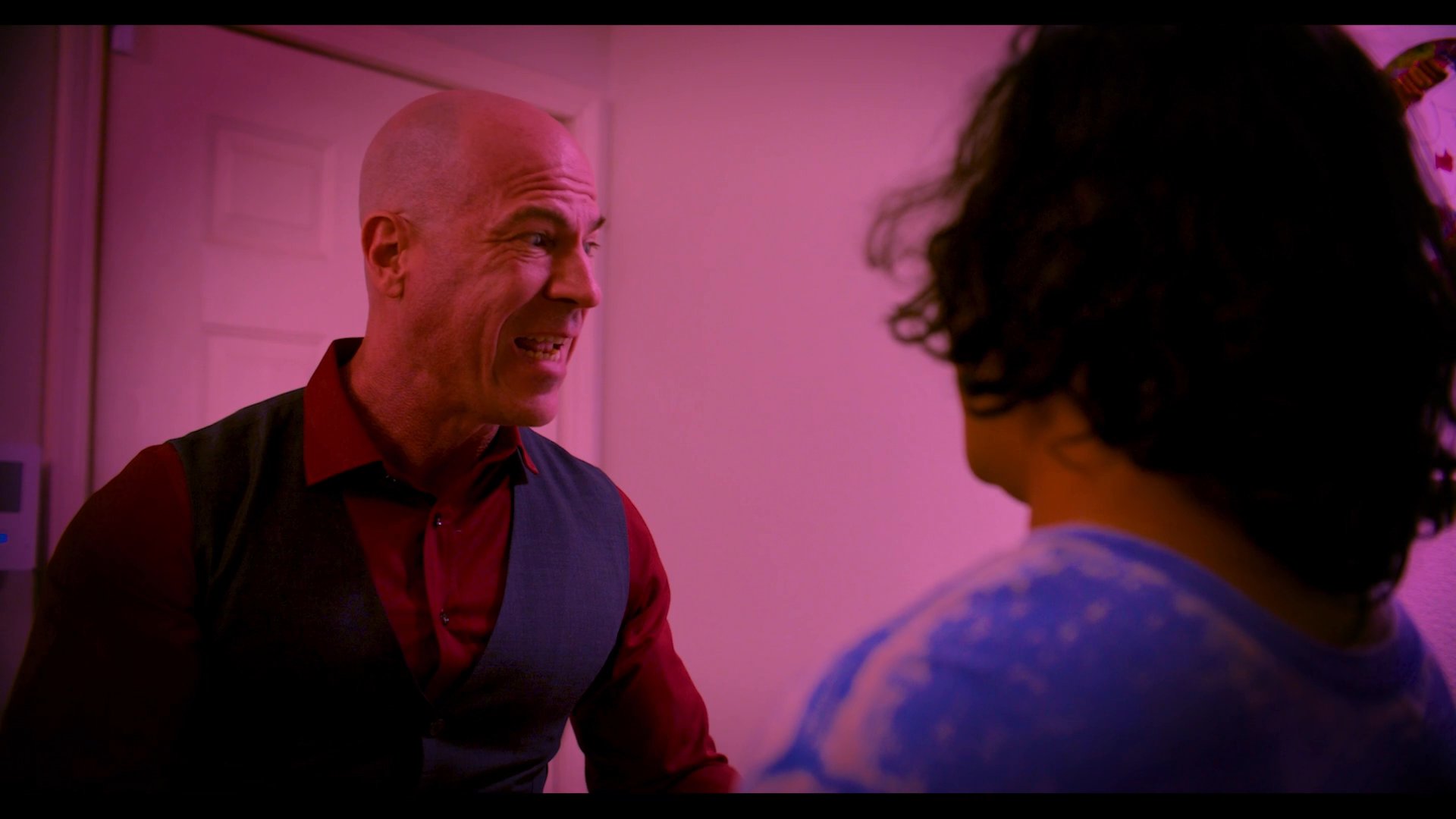 Two men engage in a tense conversation within a room featuring abstract artwork. The scene focuses on the dynamic interaction and emotional weight of their dialogue.