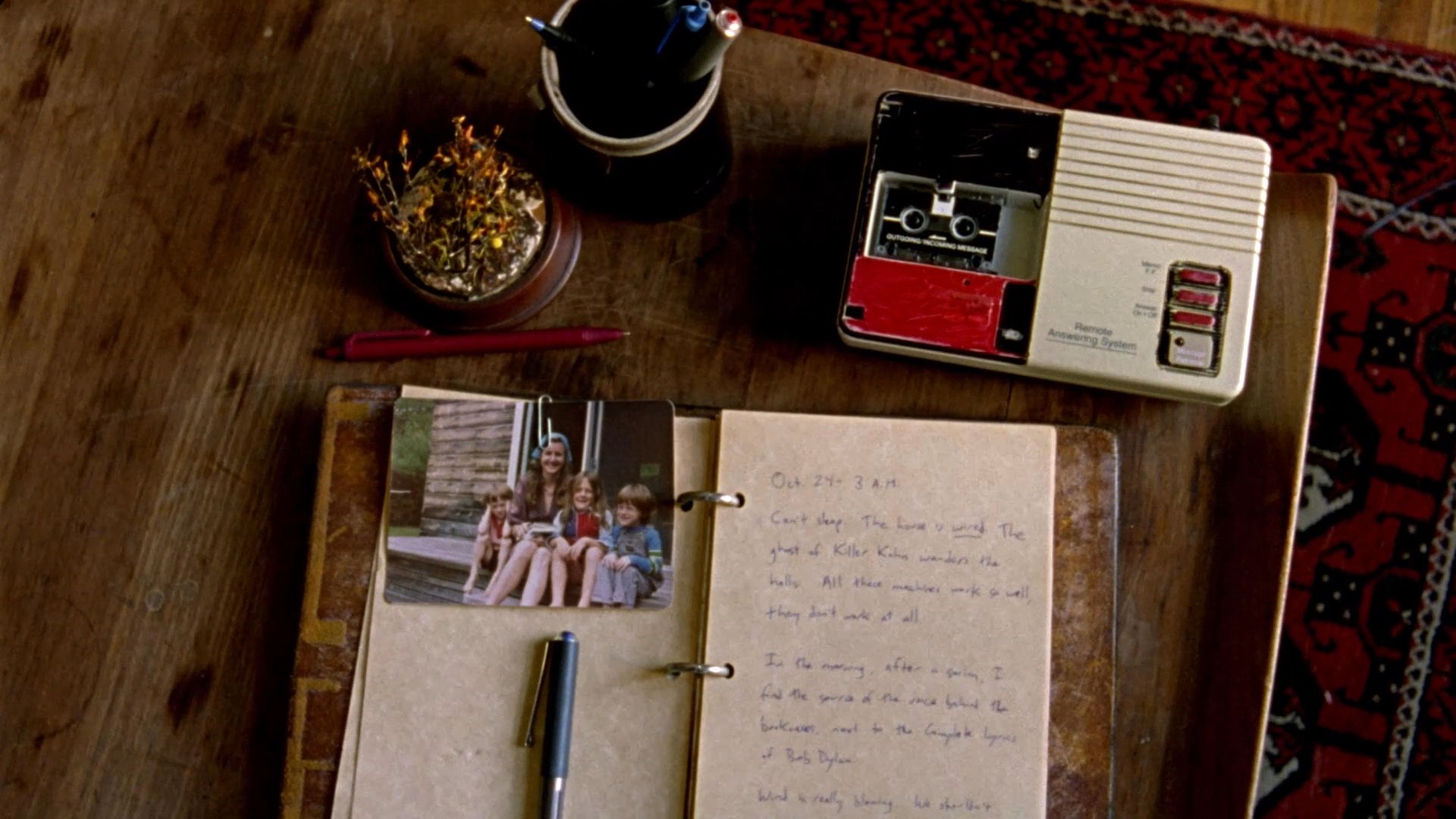 Historical news text detailing the death of Bill Graham transitions into an intimate view of a personal journal featuring a photograph and handwritten reflections. The scene captures a moment of remembrance through the examination of preserved private records.