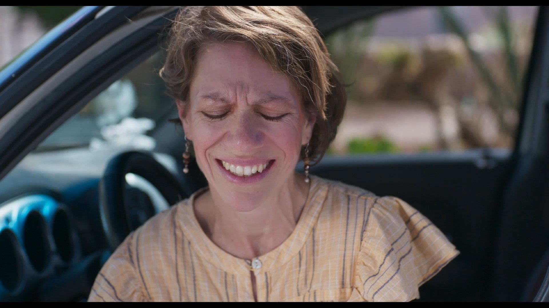 A woman sits in her car and experiences a wave of strong emotion while reading a handwritten Spanish note. She then drives away from a school building, reflecting on the message she received.