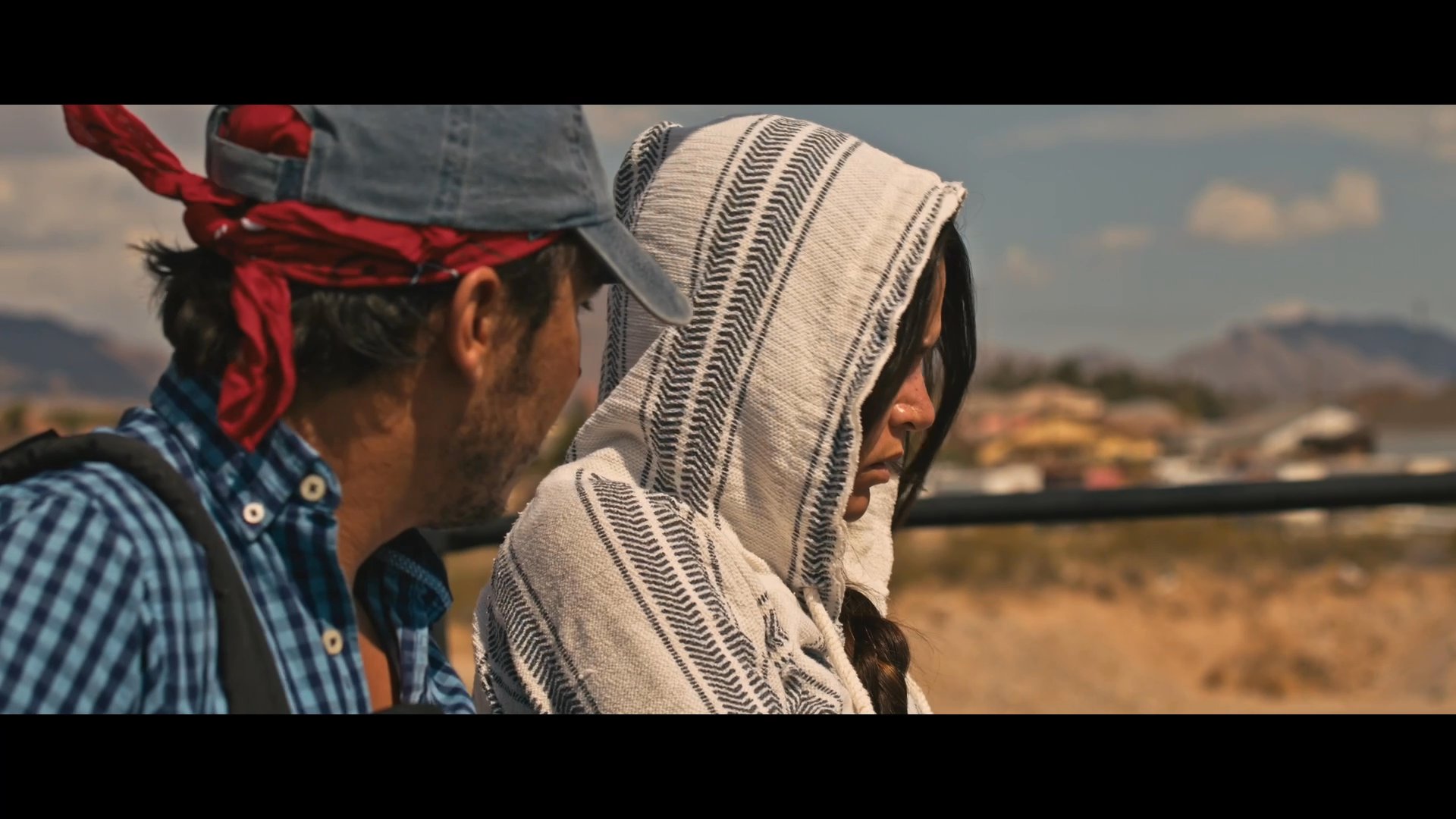Two individuals navigate the top of a moving freight train while traveling through an arid desert landscape. They carefully assist each other across the train roof, emphasizing the precarious nature of their journey.