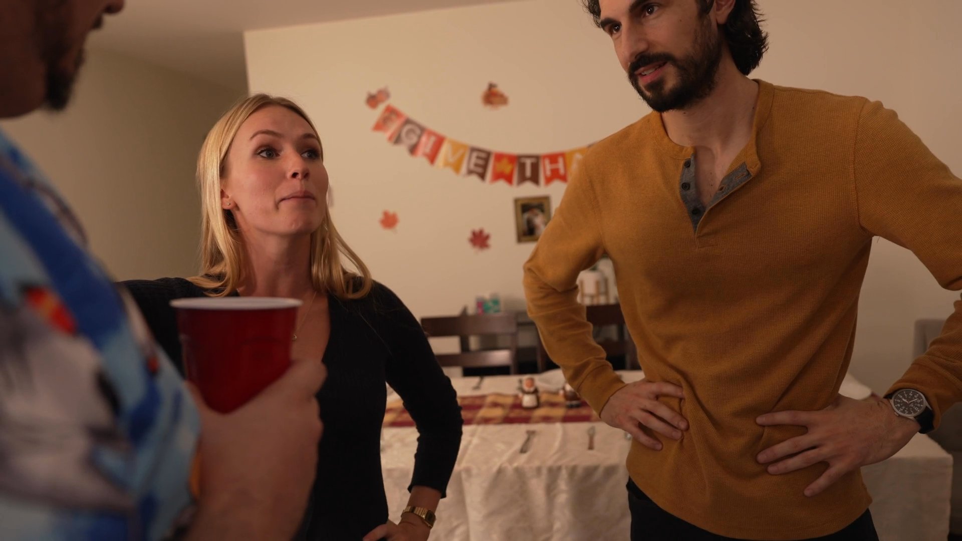 A group of friends interacts and converses in a home setting during a social gathering. The scene captures candid moments of shared laughter and casual conversation in a decorated living and dining area.
