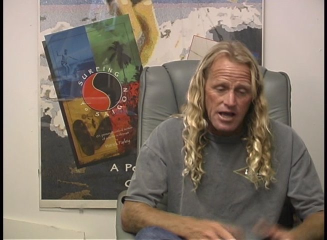 A surfer with long blonde hair discusses industry topics while seated in a studio office setting. He uses hand gestures to emphasize his points during the interview.
