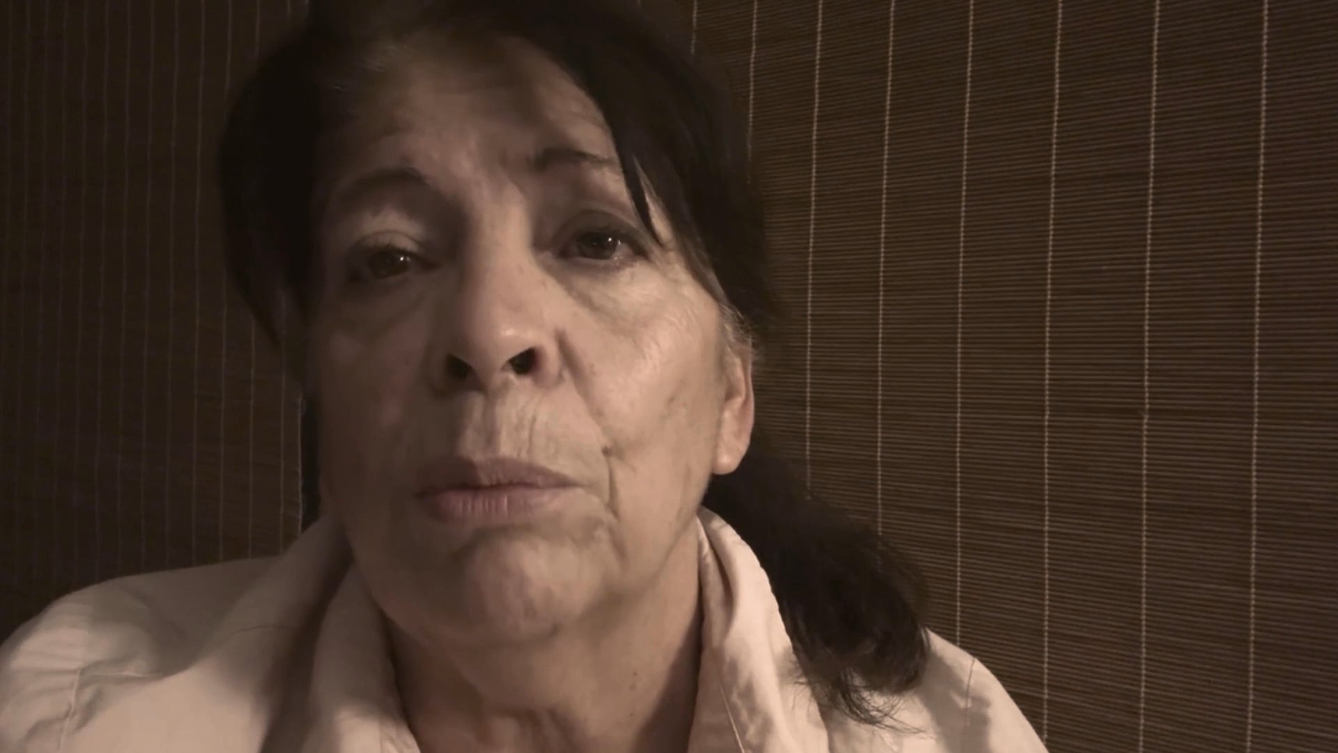 An elderly woman speaks with evident emotional distress and sorrow while sitting in a dimly lit, private space. She periodically touches her face as she shares her perspective, highlighting a moment of profound personal reflection.