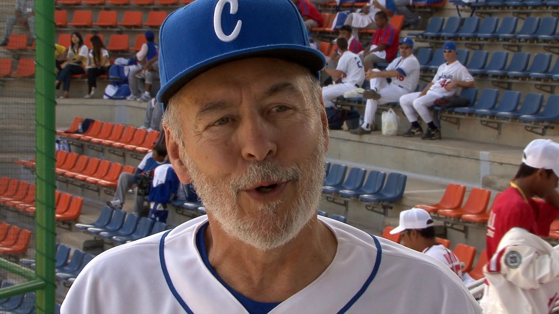 A seasoned baseball coach discusses team performance while standing in a stadium setting. Players in uniform are visible in the background as the interview takes place.