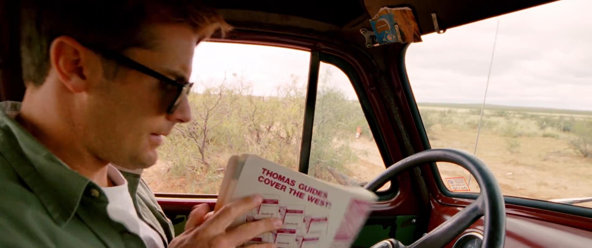 A man drives a classic truck through a remote desert landscape while consulting a paper map to navigate his route.