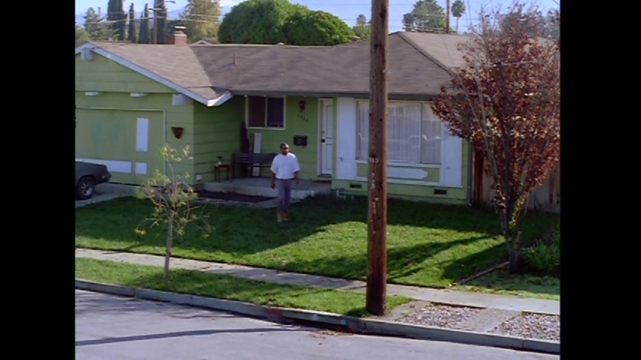 A man walks across a residential lawn and proceeds down the sidewalk in front of a green suburban house. A vehicle passes on the street during his movement.