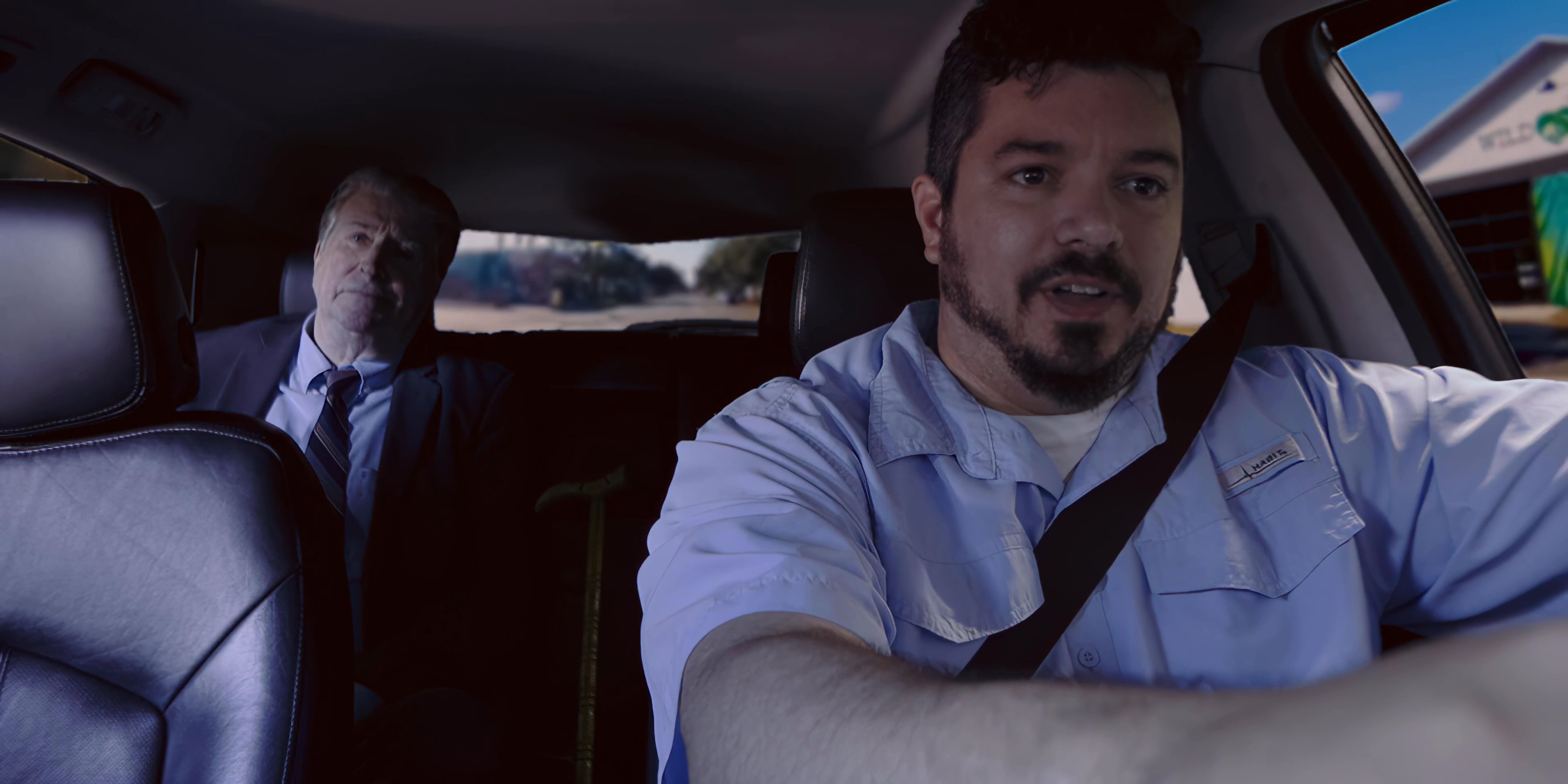 A driver and passenger engage in a serious, focused conversation while traveling inside a moving vehicle. The exchange highlights the professional or personal rapport between the two men as they navigate through a suburban setting.