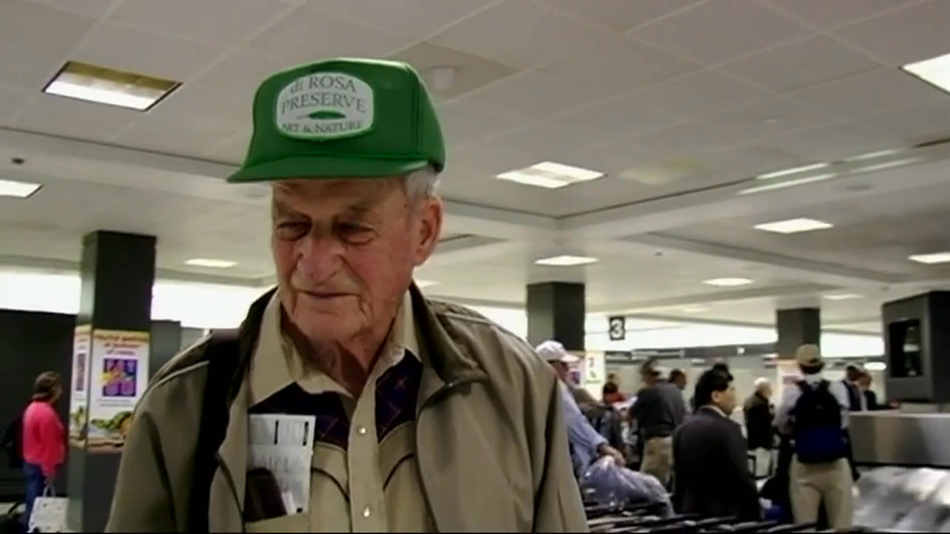 An elderly man in a green cap speaks outdoors and later inside a busy airport terminal, describing a sleepless night spent thinking about the day ahead. Shuttle buses pass beneath the Washington, D.C. airport canopy.