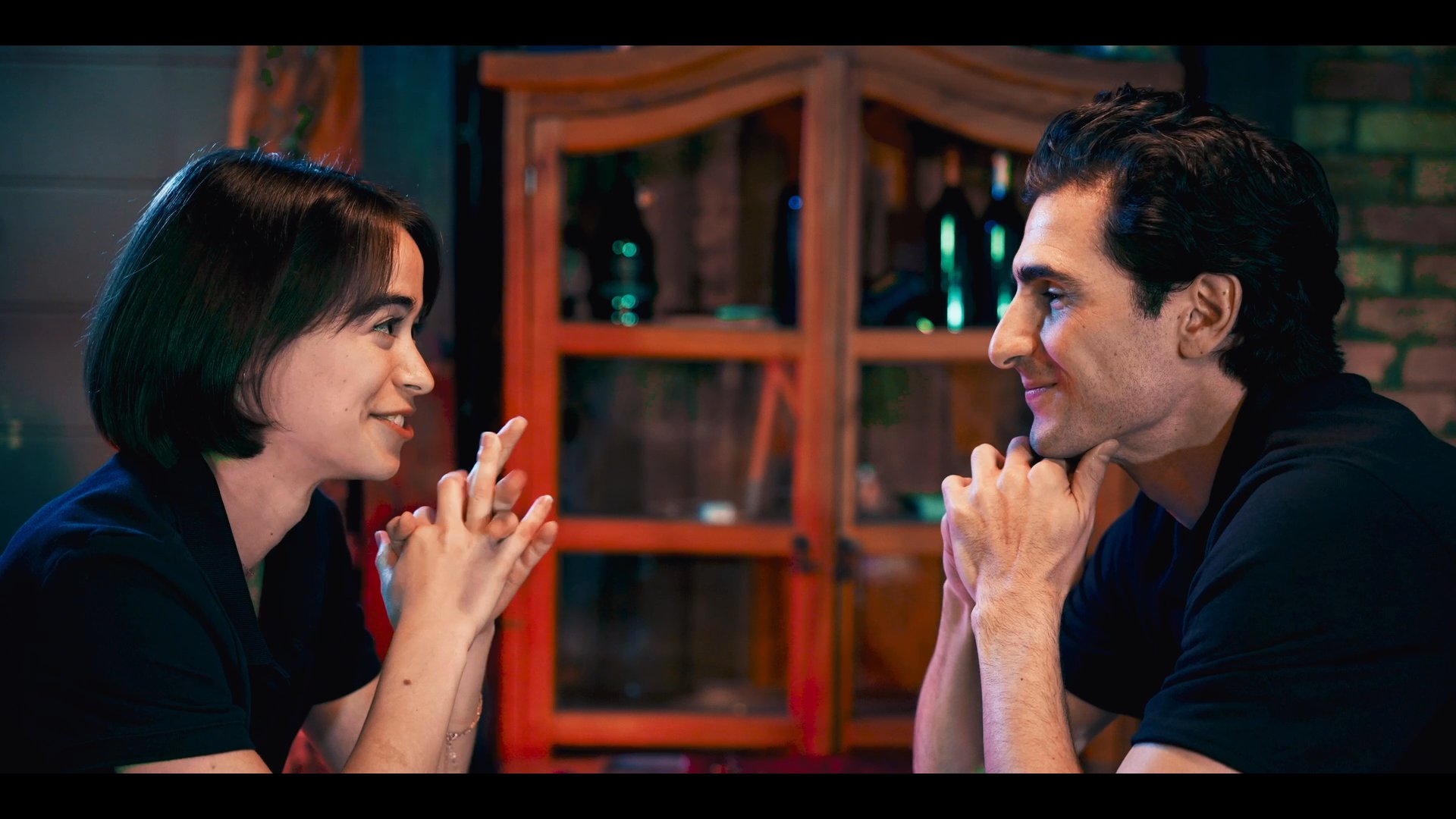 A man and woman engage in a warm conversation while seated at a bar, eventually receiving service from a bartender. The warm lighting and casual setting create an intimate social atmosphere.